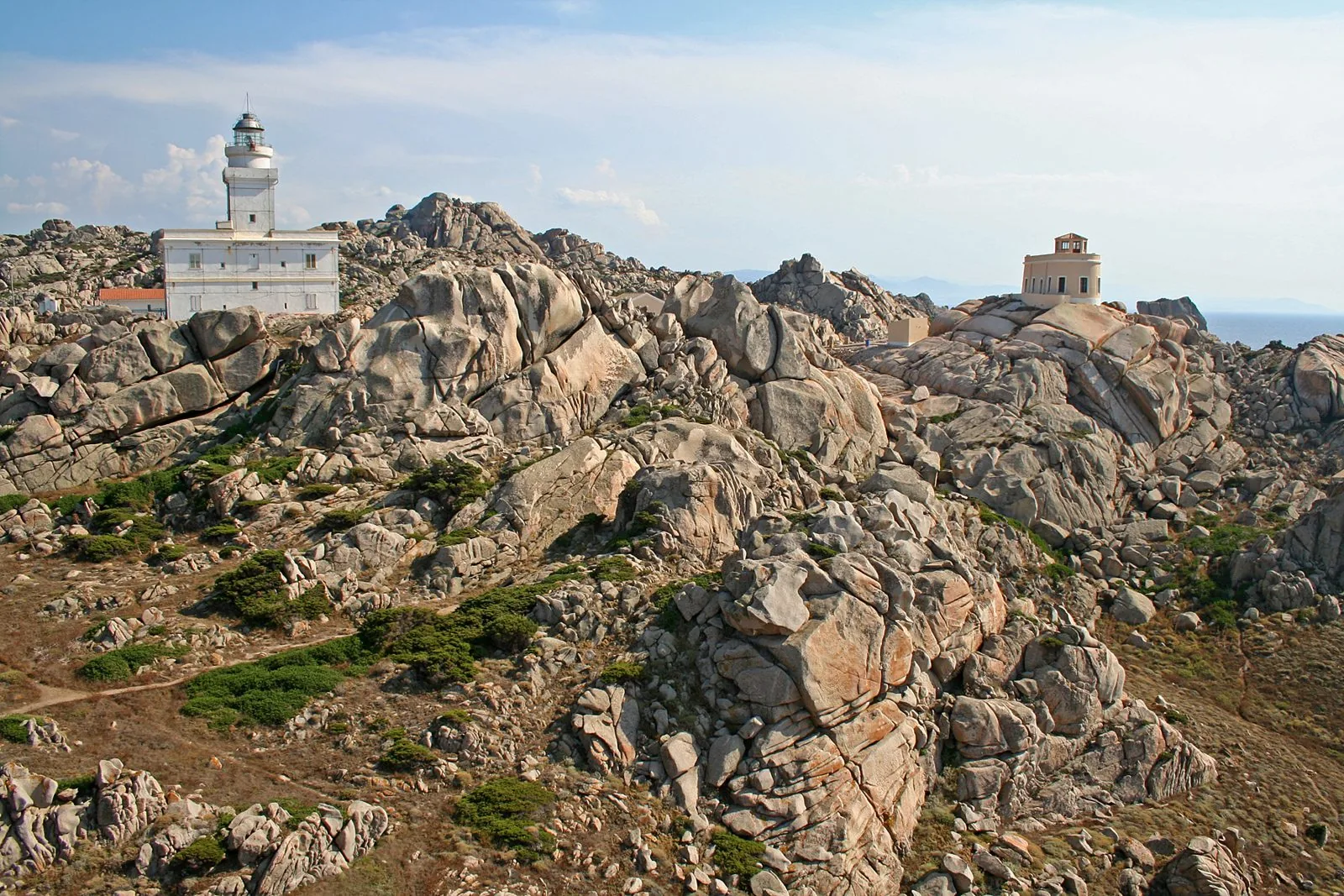 Grandi massi di granito chiaro modellati dall'erosione a Capo Testa, con cespugli di macchia e il faro bianco sullo sfondo