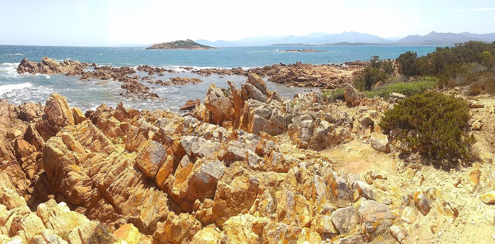 Costa di granito rosato con mare mosso e onde bianche, un isolotto e le montagne della Gallura sullo sfondo